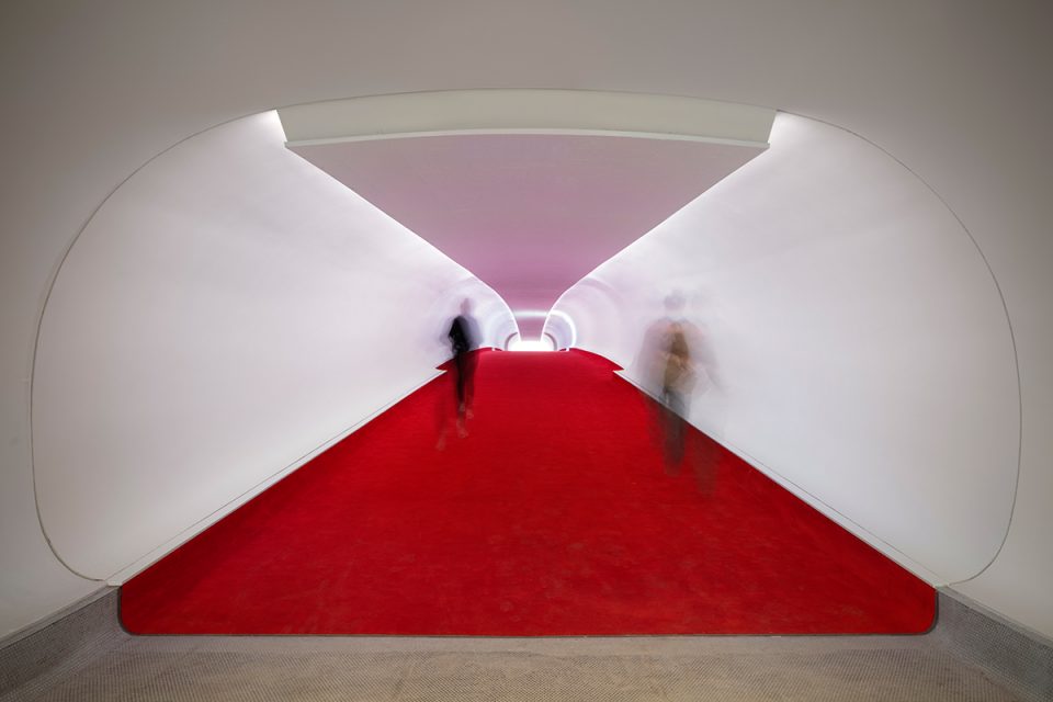 Image of red-carpet lined white tubes and blurred people walking through