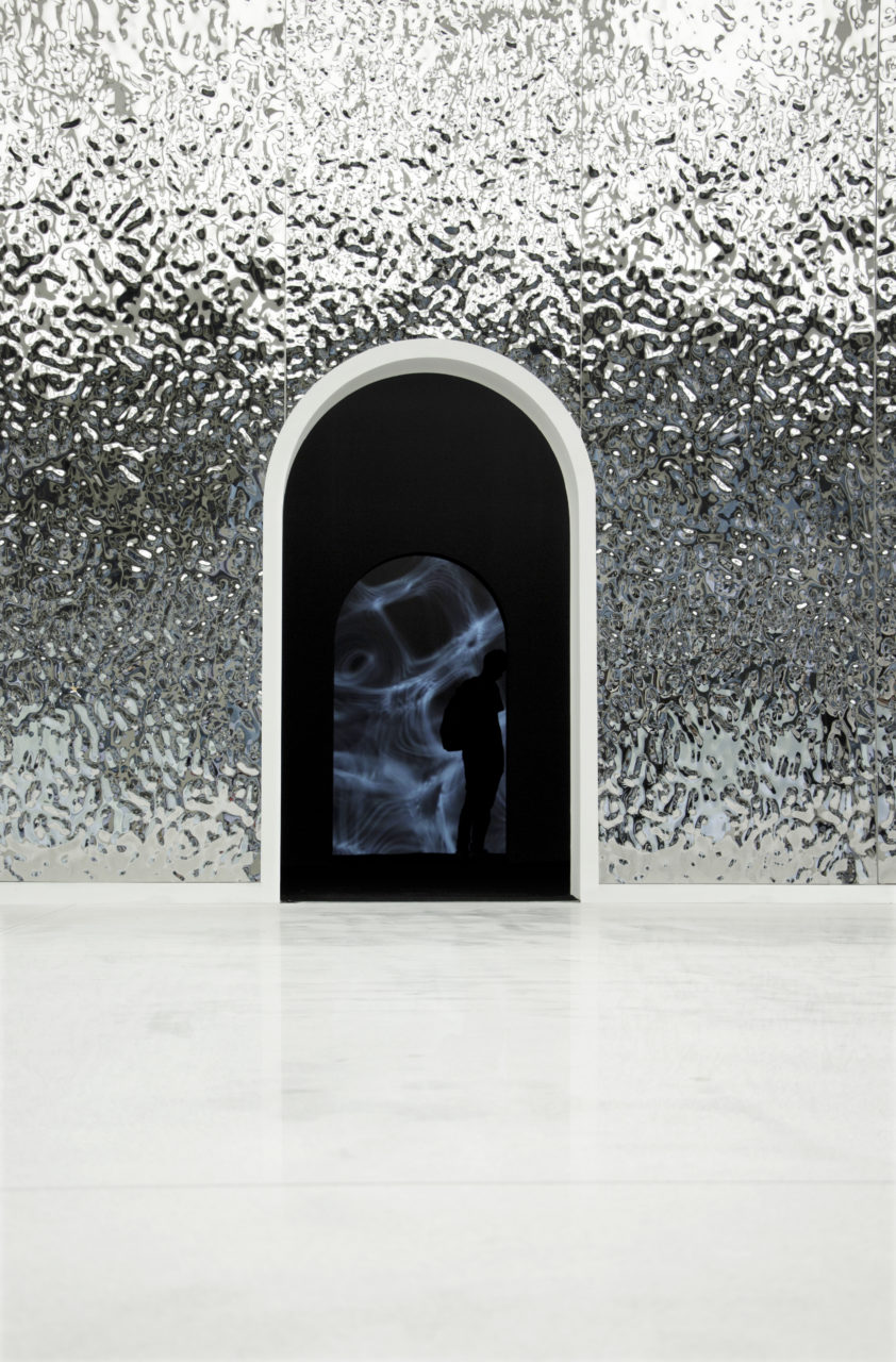 A rendering of an archway cut into a textured and reflective metallic surface. the silhouette of a figure can be found with in the archway