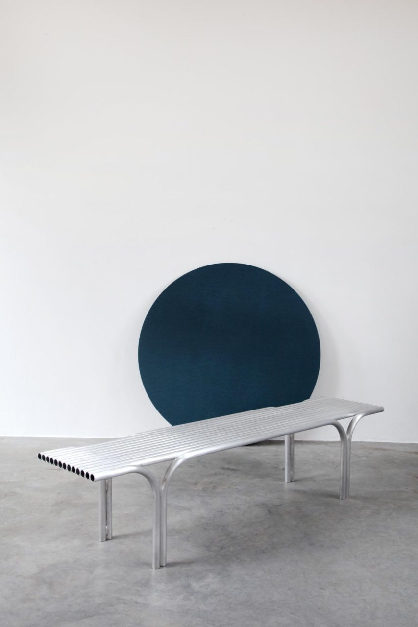 Image of a bench made of metal tubes in front of a blue circle carpet mounted on a white wall