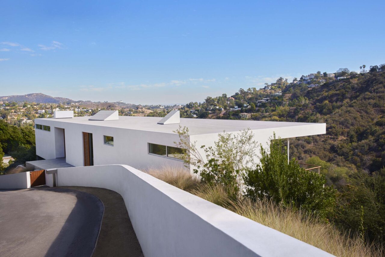 A road winds down before branching off away from a white modern, linear ranch-style home