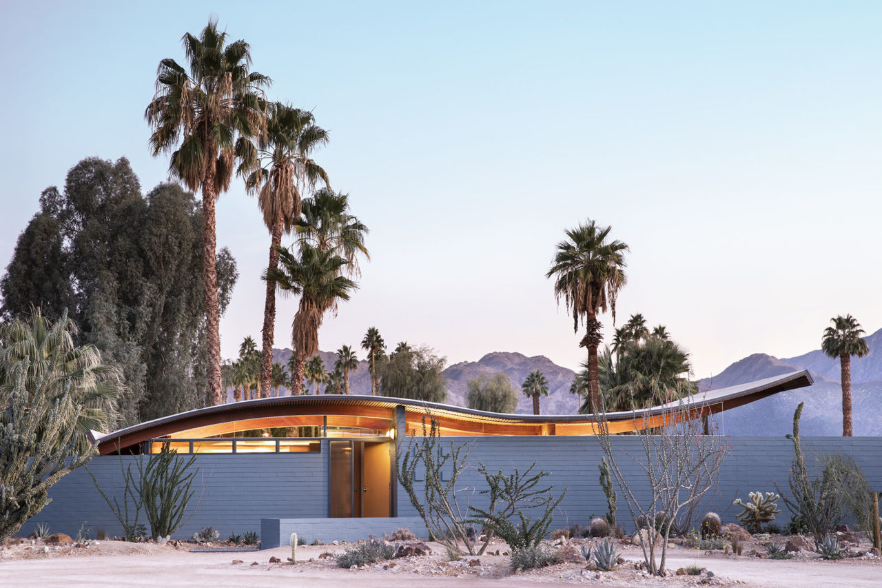 exterior view of a single story home with wave-shaped roof set in a desert landscape with palm trees