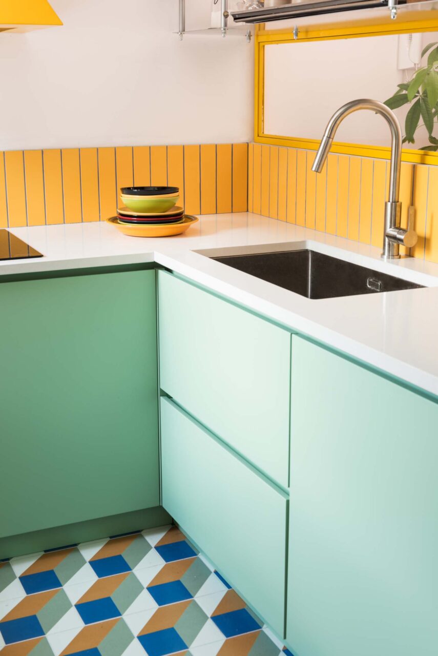 A geometric floor pattern, teal cabinets, and yellow cladding make up a small kitchen by 02A