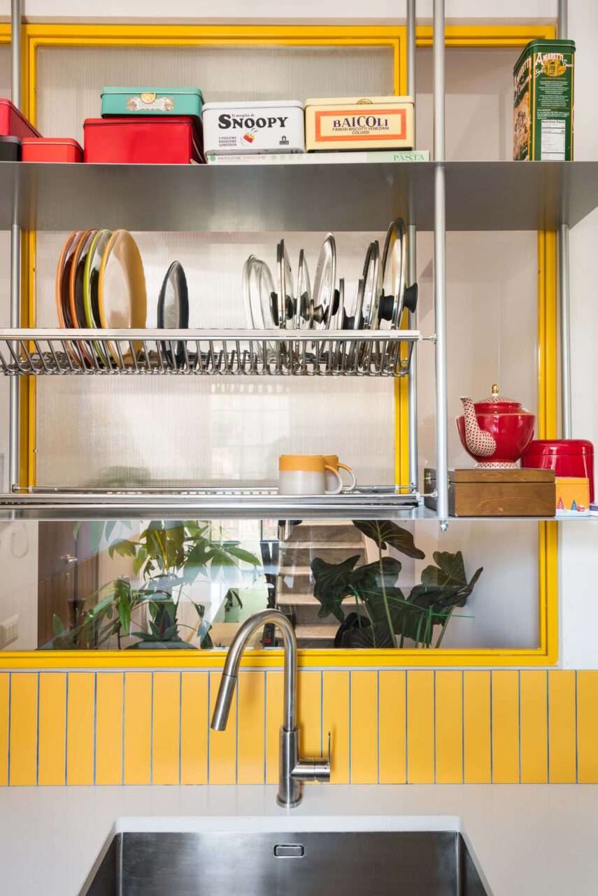 A yellow wall features metal shelving with a dish rack over the sink