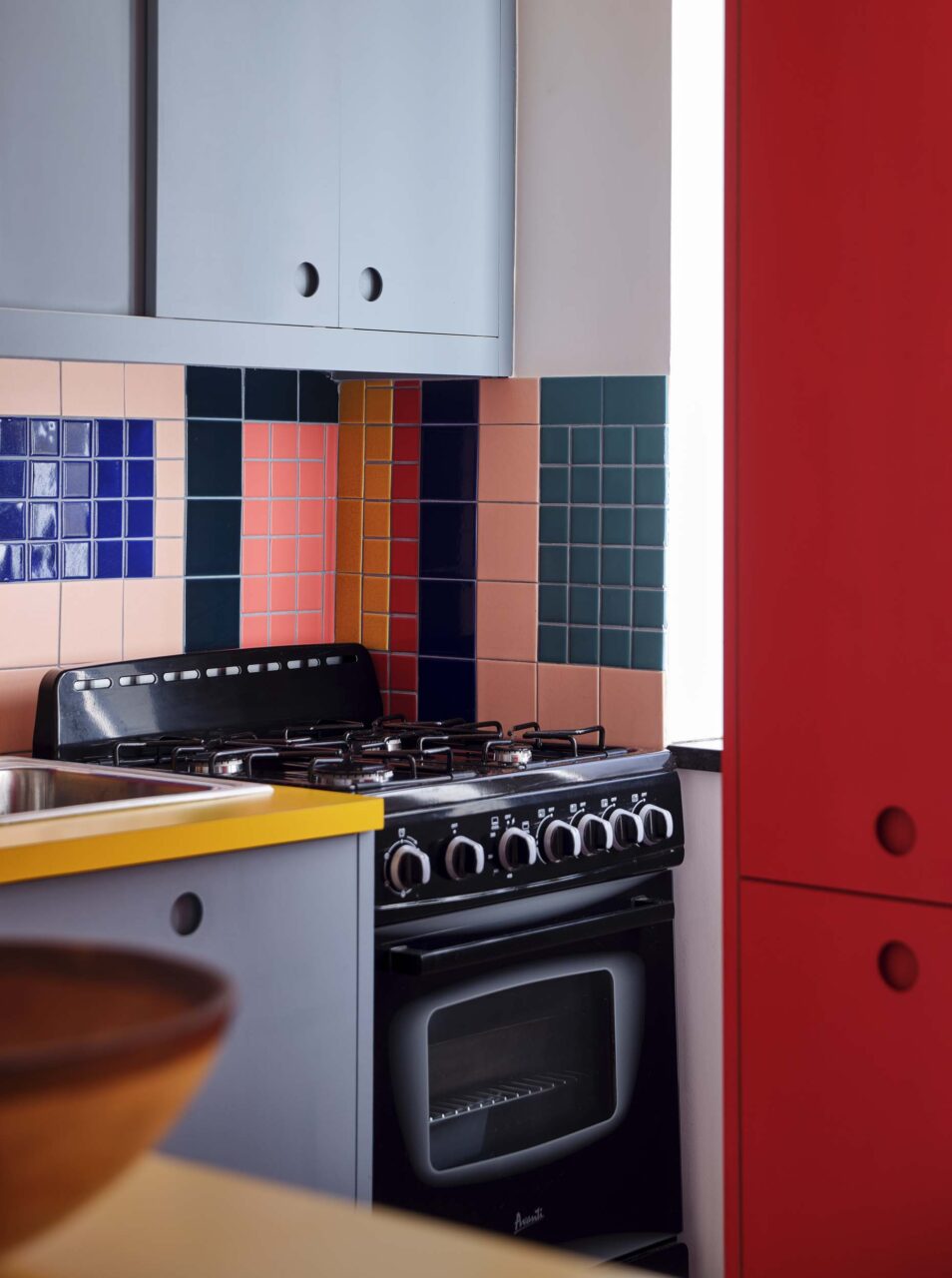 A stove is surrounded by blue, red, and yellow cabinets and counters designed by Sarah Jacoby